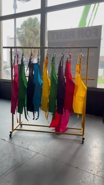 A rolling rack of brightly colored aprons — pink, green, blue, yellow, red — waiting by the studio window.